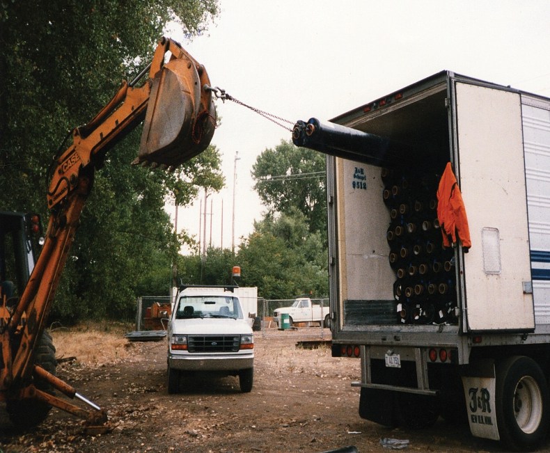 RP3XL in use — excavator arm holding a Roll Puller chained to a geosynthetic roll being pulled from the back of a J&R box truck